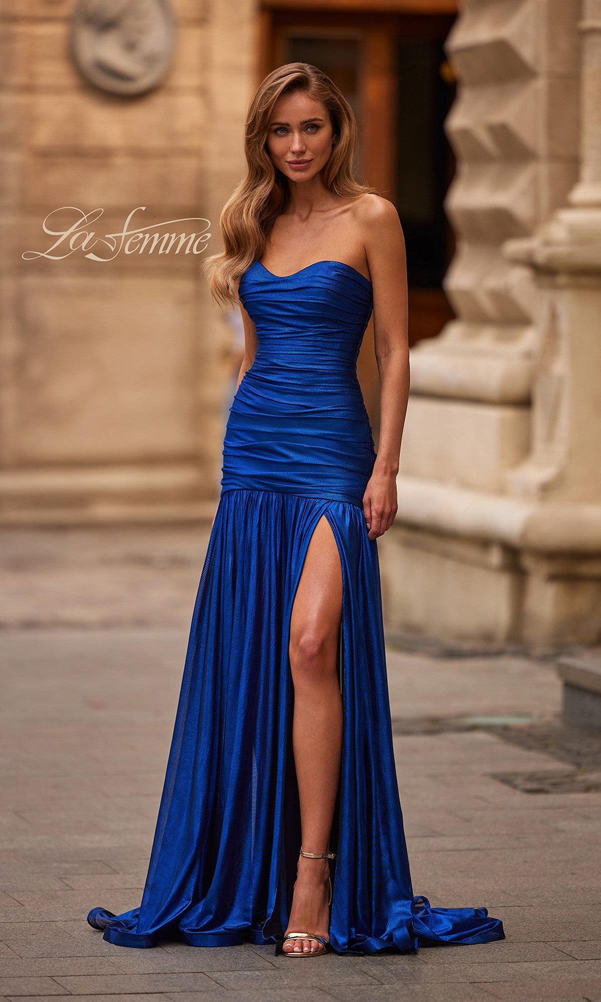 A woman wears the La Femme 33200 long prom dress, a strapless blue gown with a ruched sweetheart neckline and thigh-high slit, standing by a stone building on a city sidewalk.